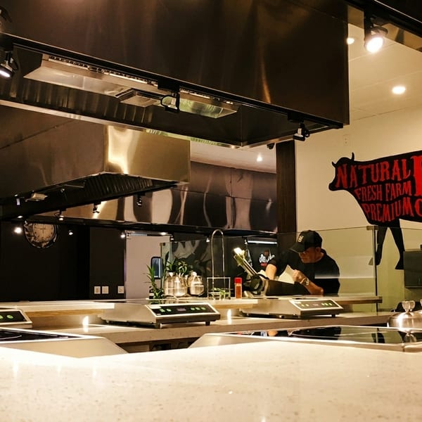 A restaurant kitchen with stainless steel hoods, cooking stations, and a "Natural Beef" sign.